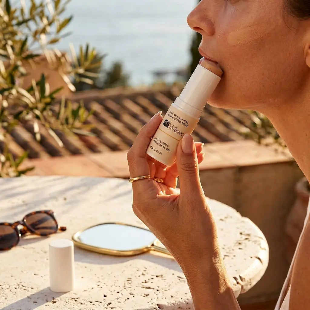 Stick de protection solaire SPF50 teinté en action: femme appliquant le produit sur les lèvres lors d'une pause en plein air, avec lunettes et miroir sur une table.