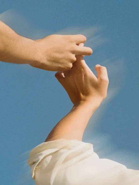 Main sujet: deux mains qui se rejoignent dans le ciel, l’une saisissant légèrement l’autre, bras nu et chemise blanche en bas. Alt: "Deux mains se connectant dans le ciel bleu, symbolisant protection et soin solaire."