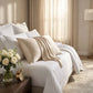Taie d’oreiller en satin clair assortie à un lit blanc, posé près d’une table de nuit avec une fleuriste et un réveil doré. Valentine.