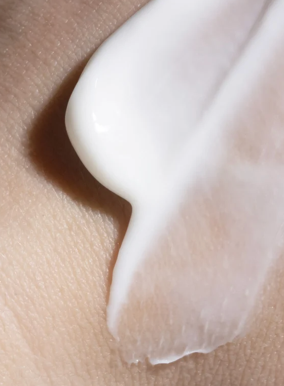 Crème corporelle blanche déposée sur la peau, texture lisse et crémeuse, avec une légère courbe. Autour, peau légèrement rugueuse.