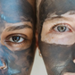 Deux visages encadrés, masqués de pâte d’argile bleu et noire couvrant la peau, yeux visibles au centre.